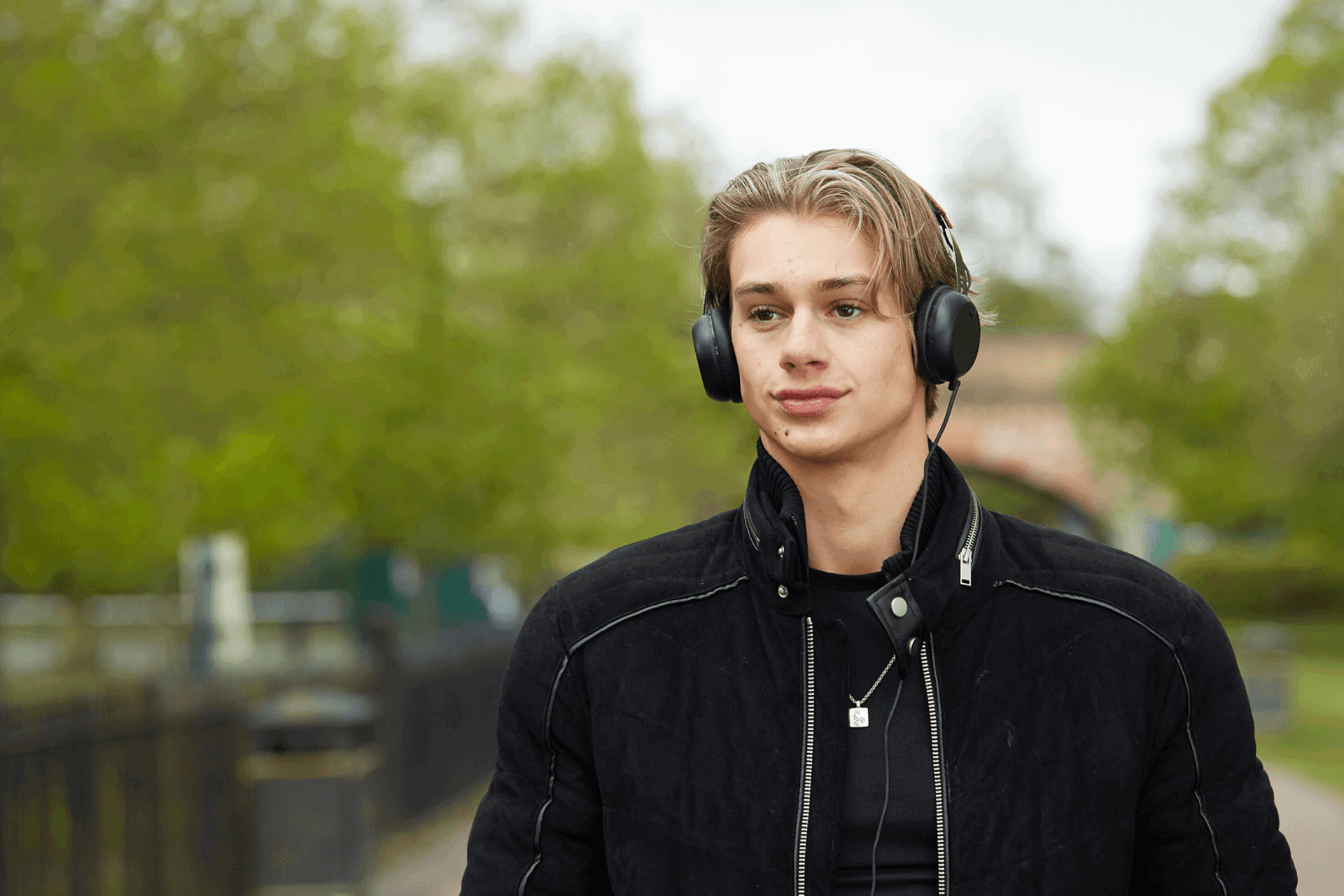 a boy wearing black jacket and with headphones on while walking on a tree lined street