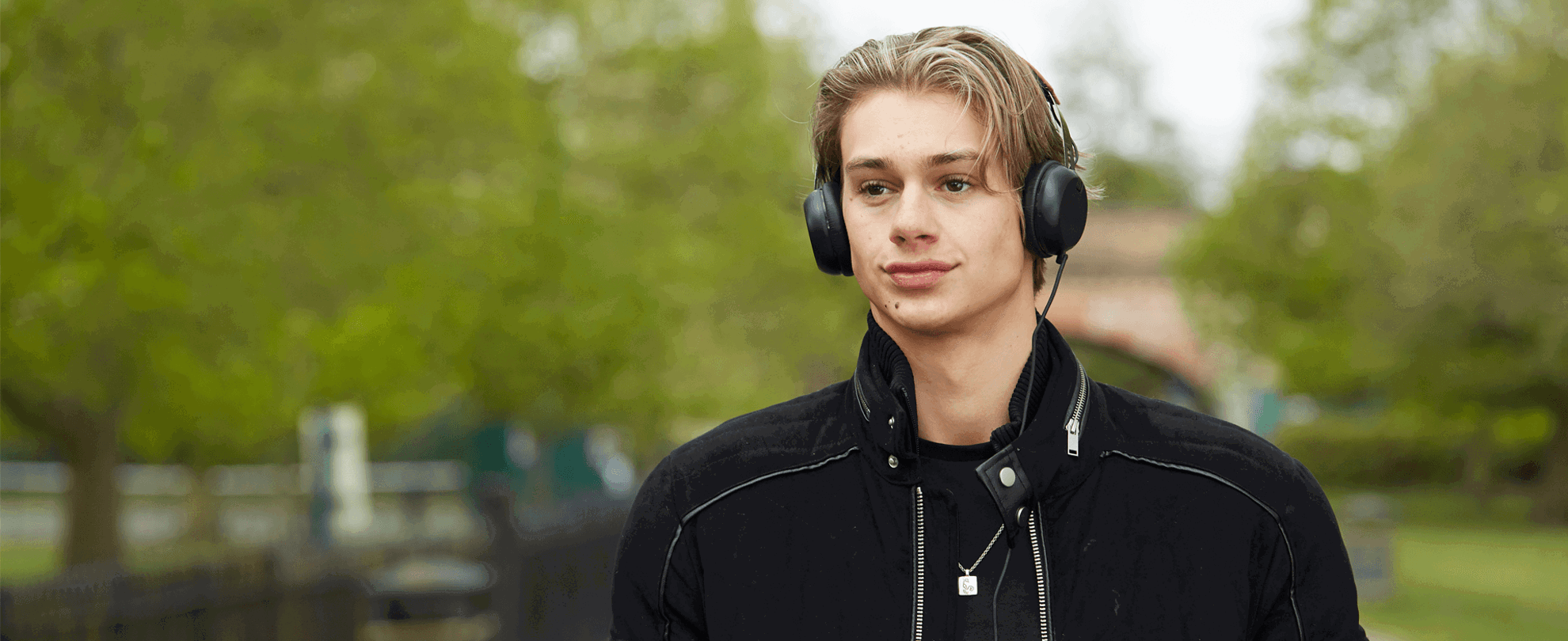 a boy wearing black jacket and with headphones on while walking on a tree lined street