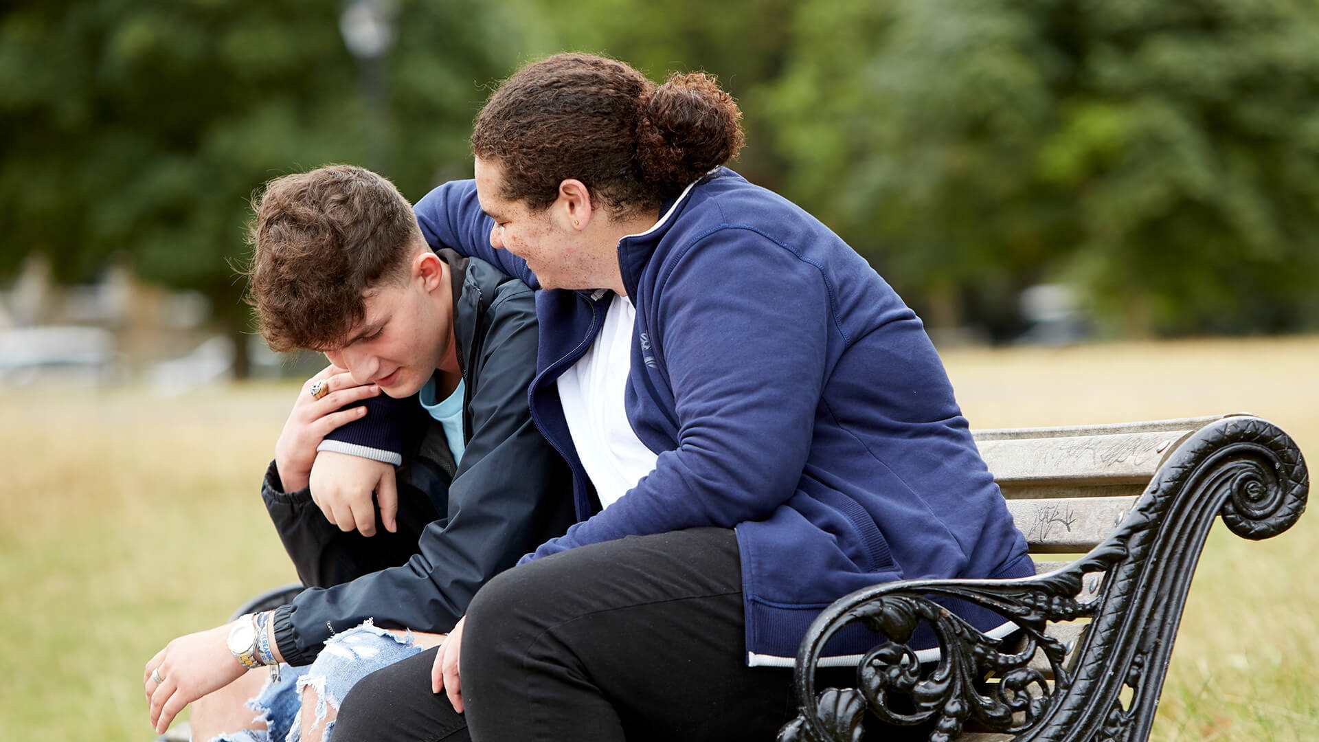 Two young people sit on a bench in a park. The person on the right has his arm around the other young person. The young person on the left is holding the other persons arm while looking down at the floor.