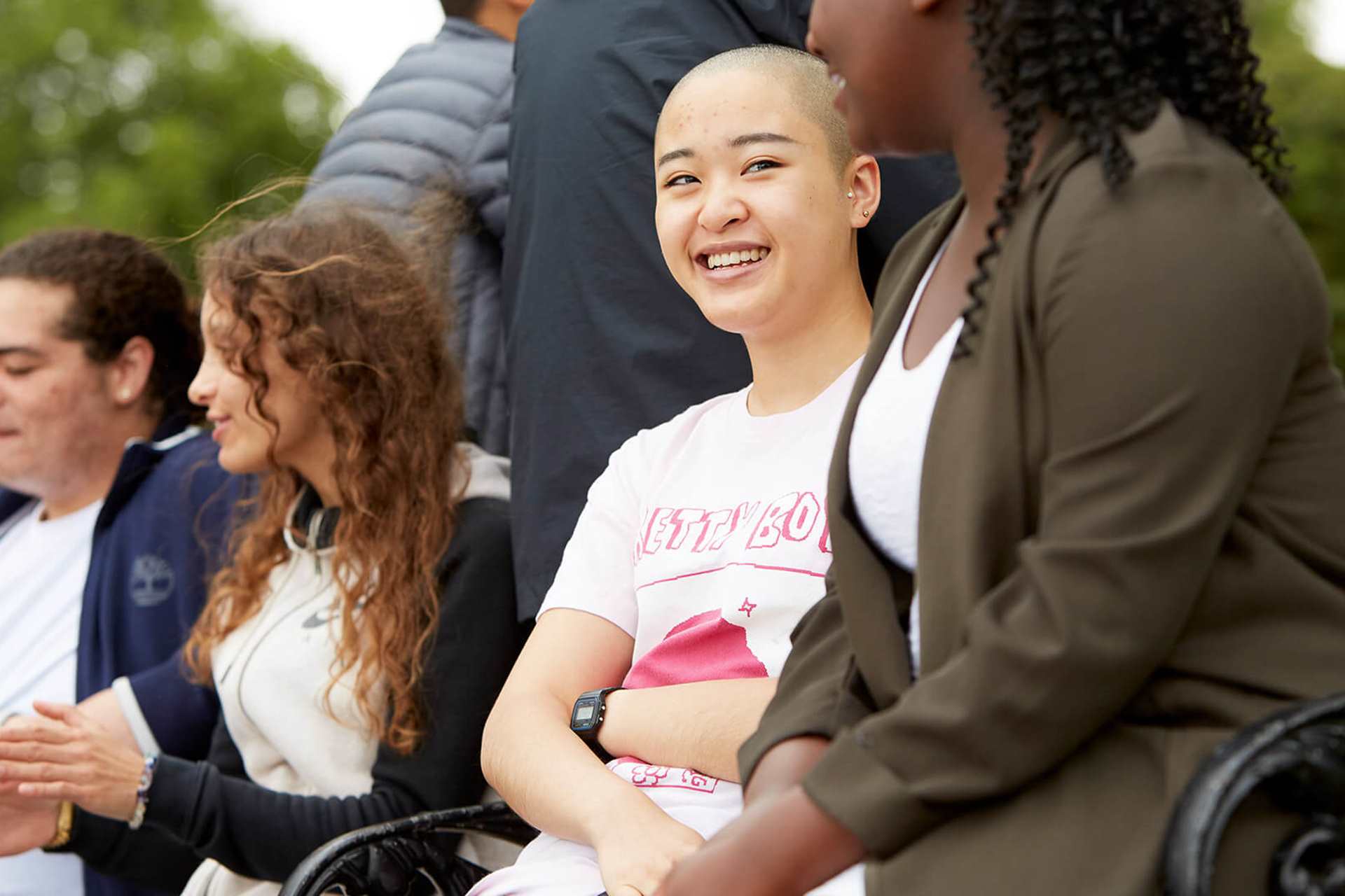 A group of young people sit on a picnic bench together in the park. They are smiling and talking together.