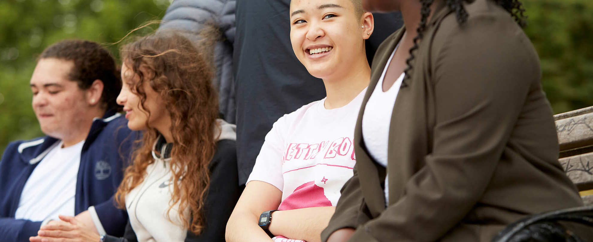A group of young people sit on a picnic bench together in the park. They are smiling and talking together.