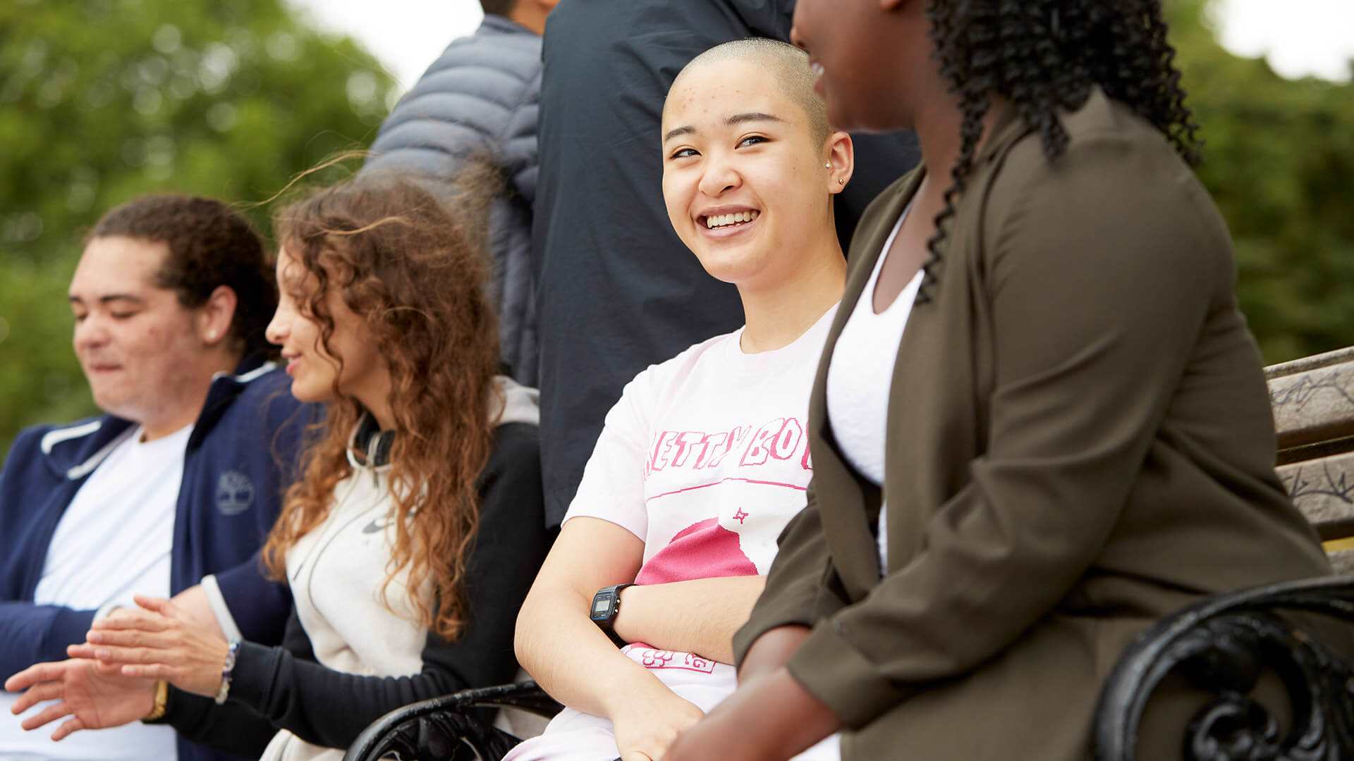 A group of young people sit on a picnic bench together in the park. They are smiling and talking together.