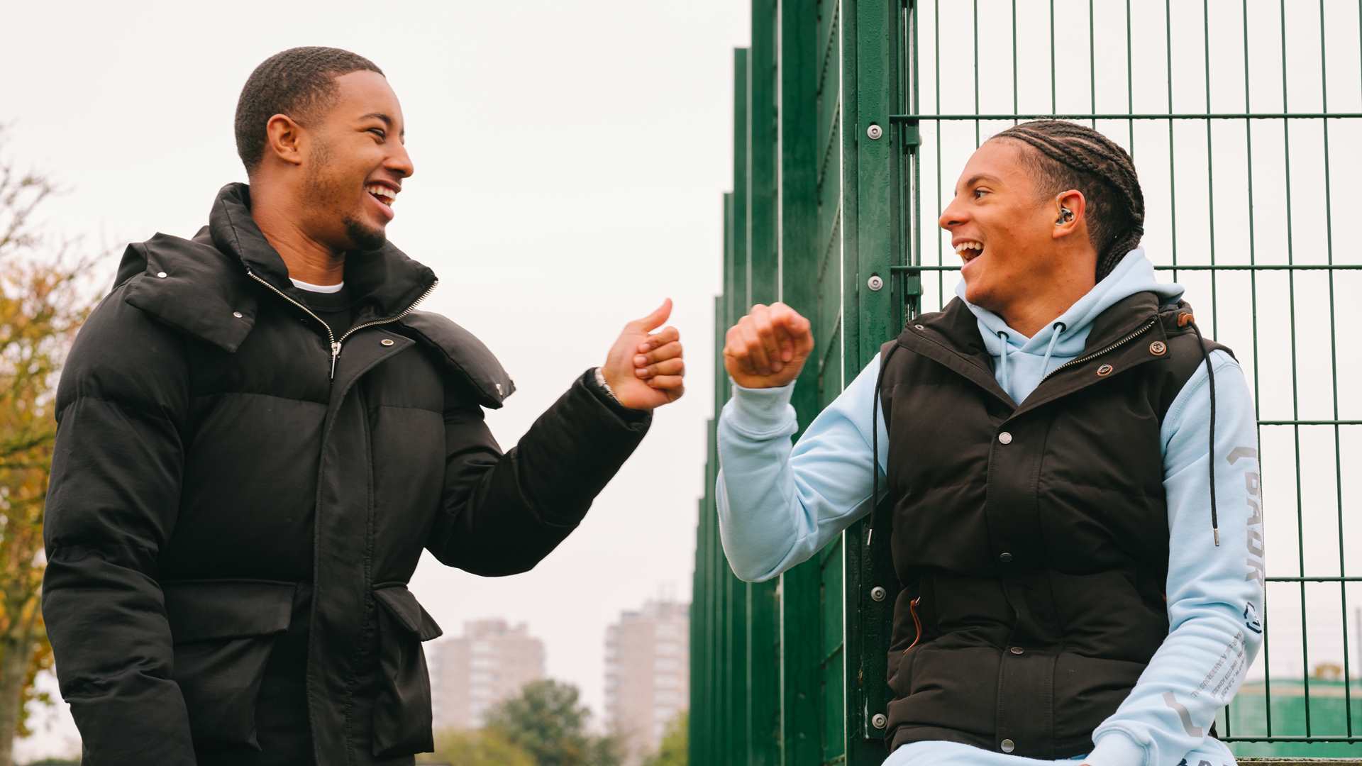 A Black teenage boy wearing a hearing aid bumping fists with a young Black man in the park.