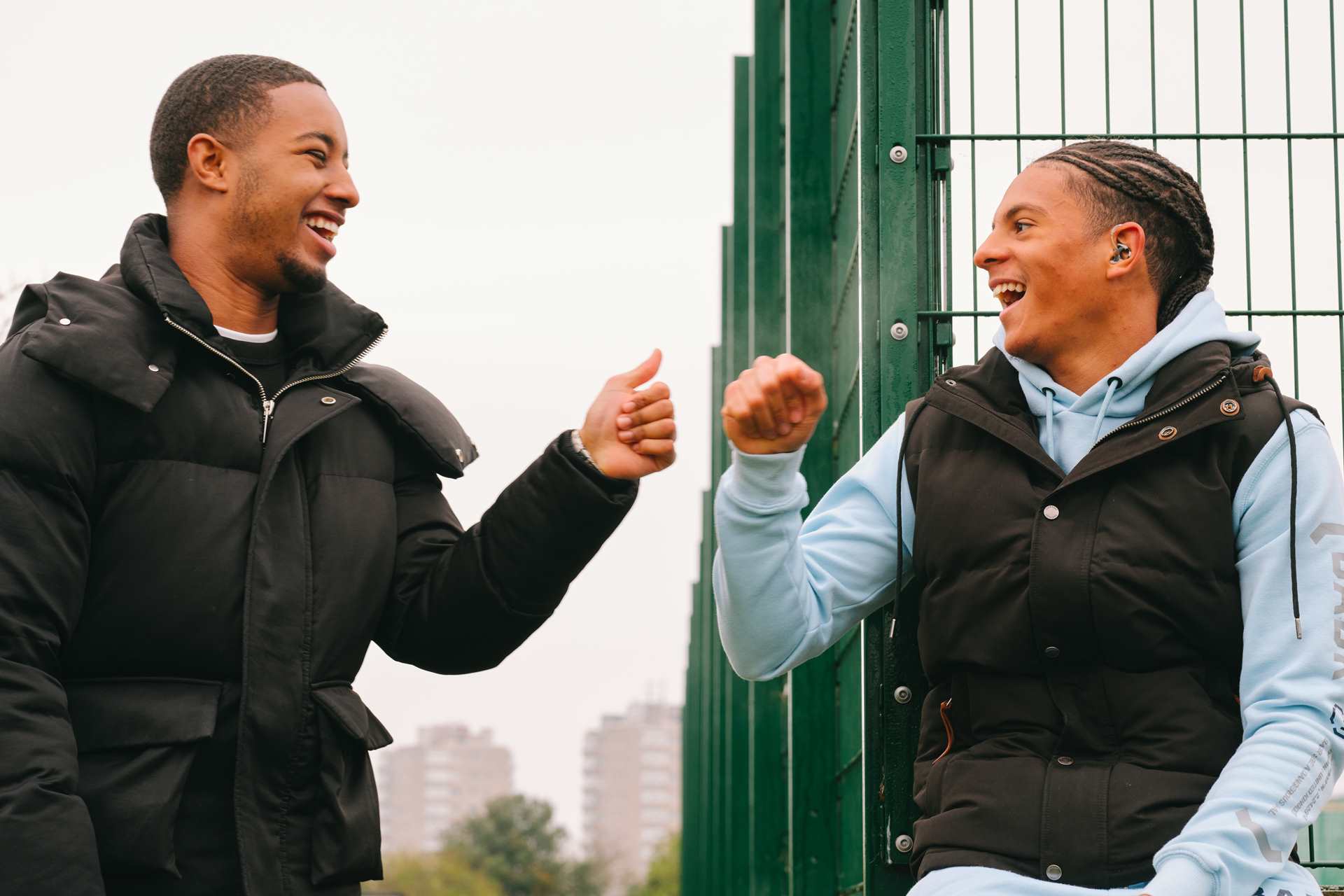 A Black teenage boy wearing a hearing aid bumping fists with a young Black man in the park.