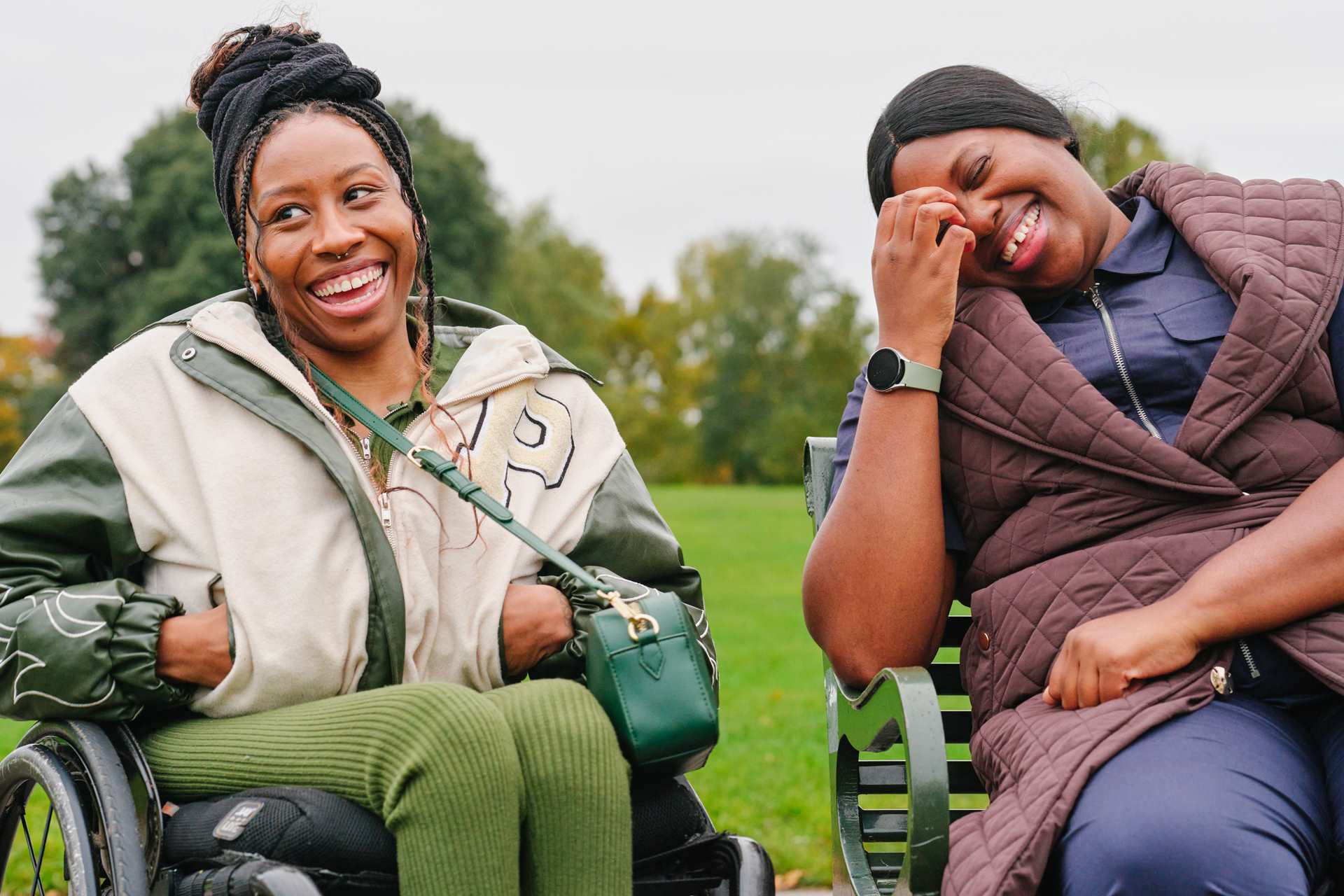 A young Black woman in a wheelchair and an older Black woman sitting on a bench in the park. They are laughing together.