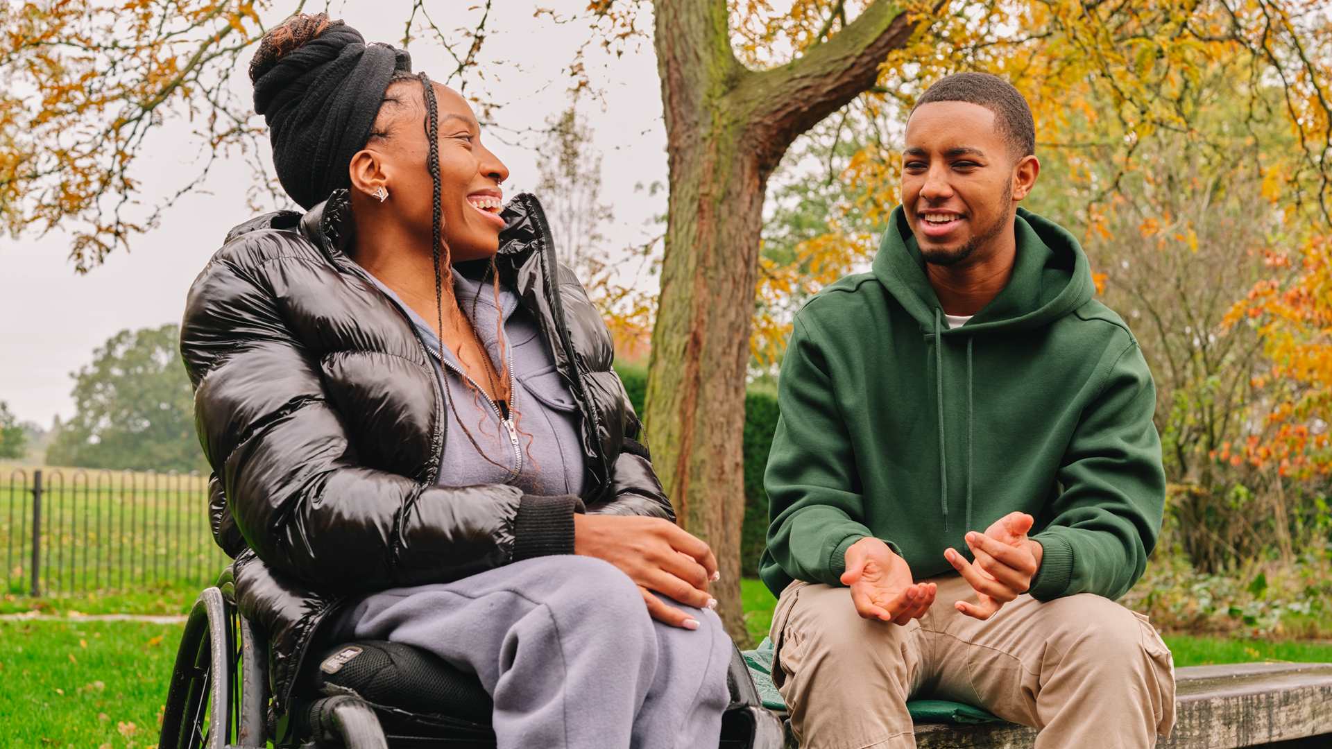 A young Black woman in a wheelchair talking to a young Black man on a bench in the park. The woman is laughing while the man explains something.