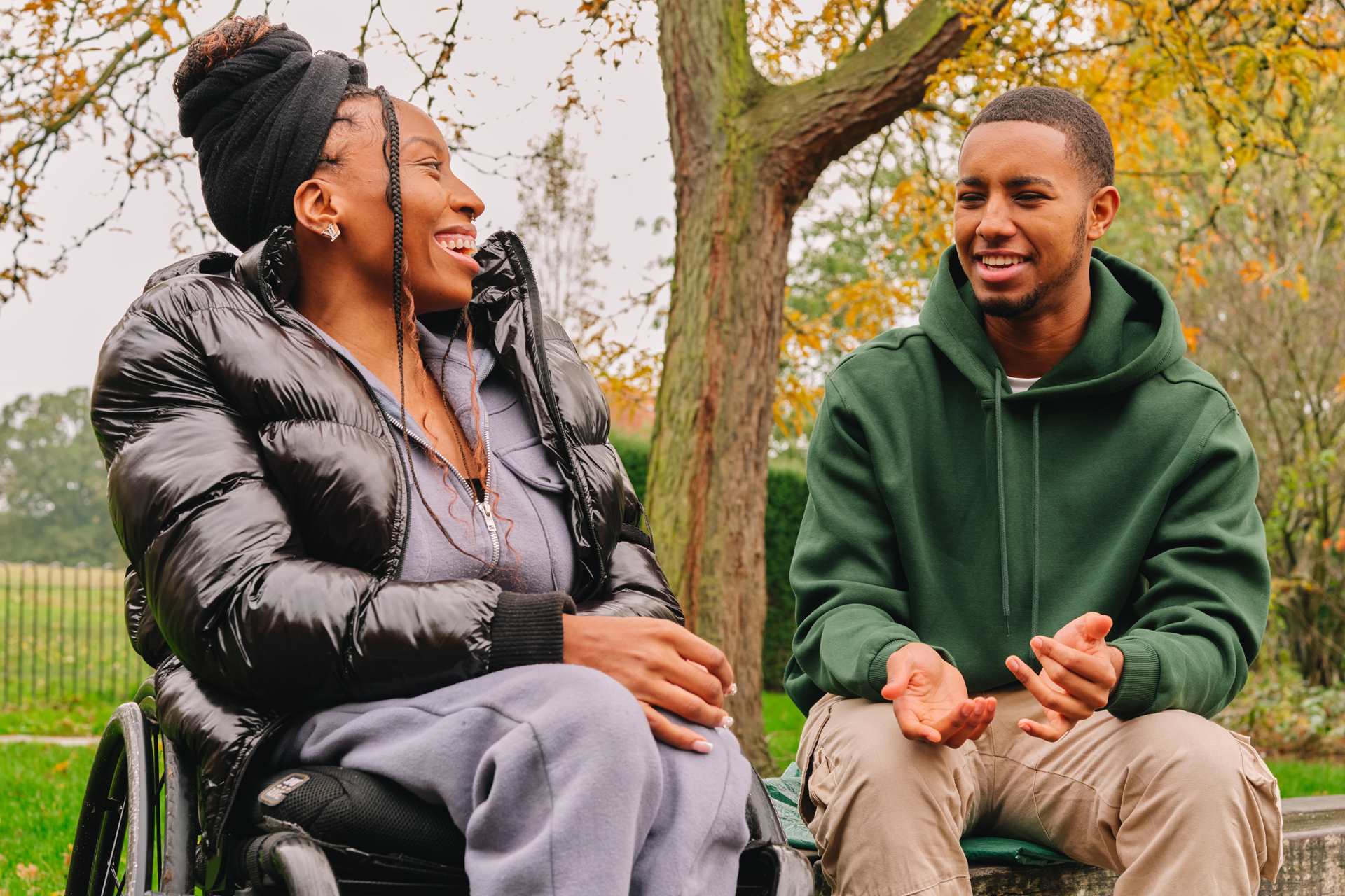 A young Black woman in a wheelchair talking to a young Black man on a bench in the park. The woman is laughing while the man explains something.