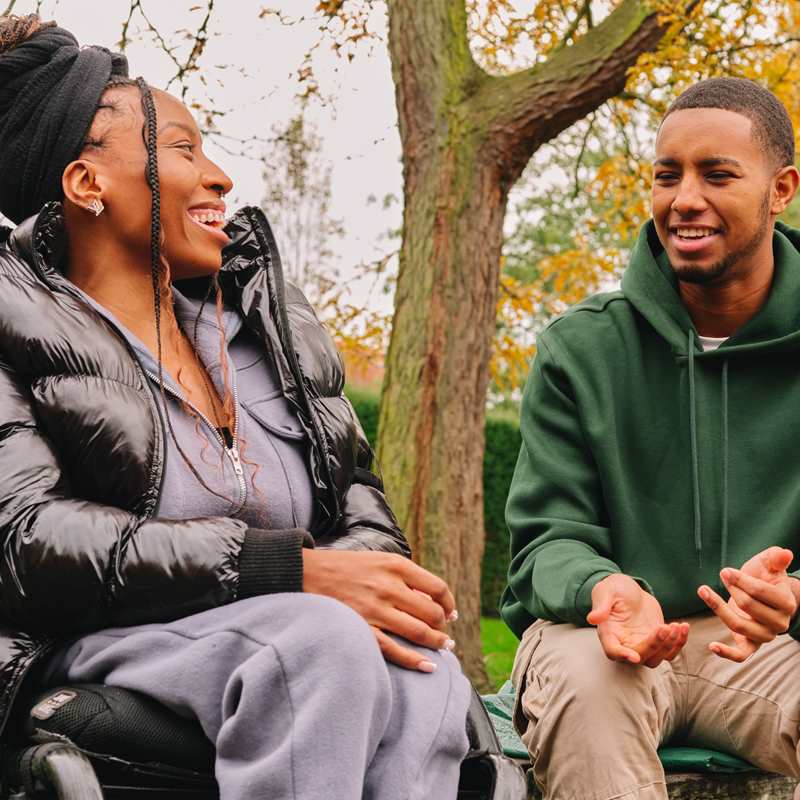 A young Black woman in a wheelchair talking to a young Black man on a bench in the park. The woman is laughing while the man explains something.