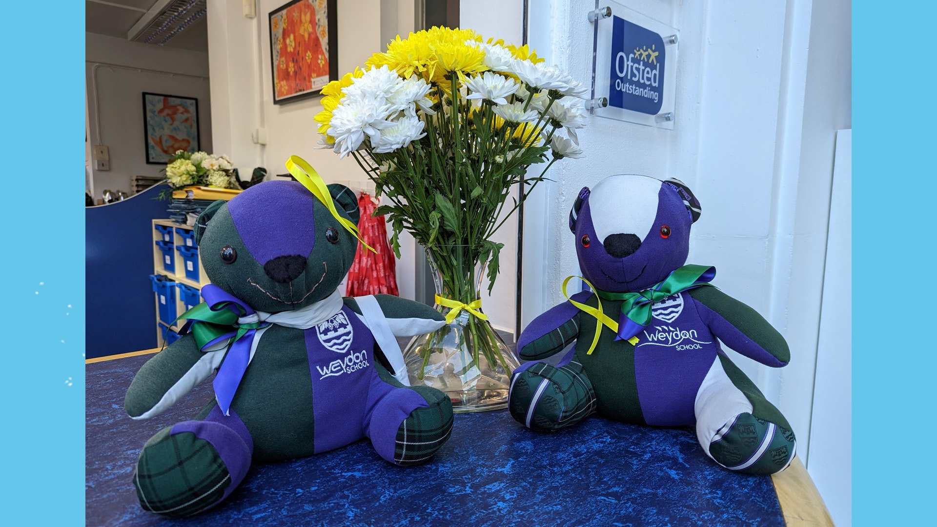 Two blue and green teddy bears on a table, with a vase of yellow and white flowers between them.