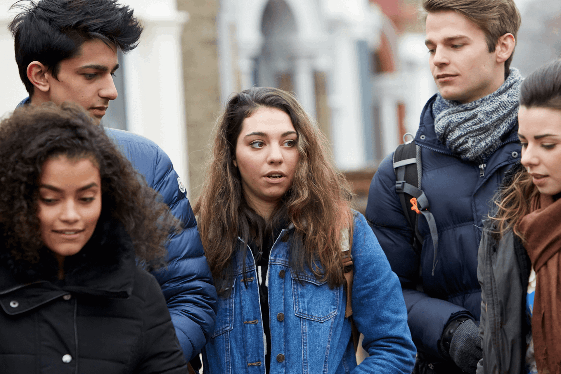 Five young people wearing coats, jackets and backpacks. They are talking as they walk down a street.