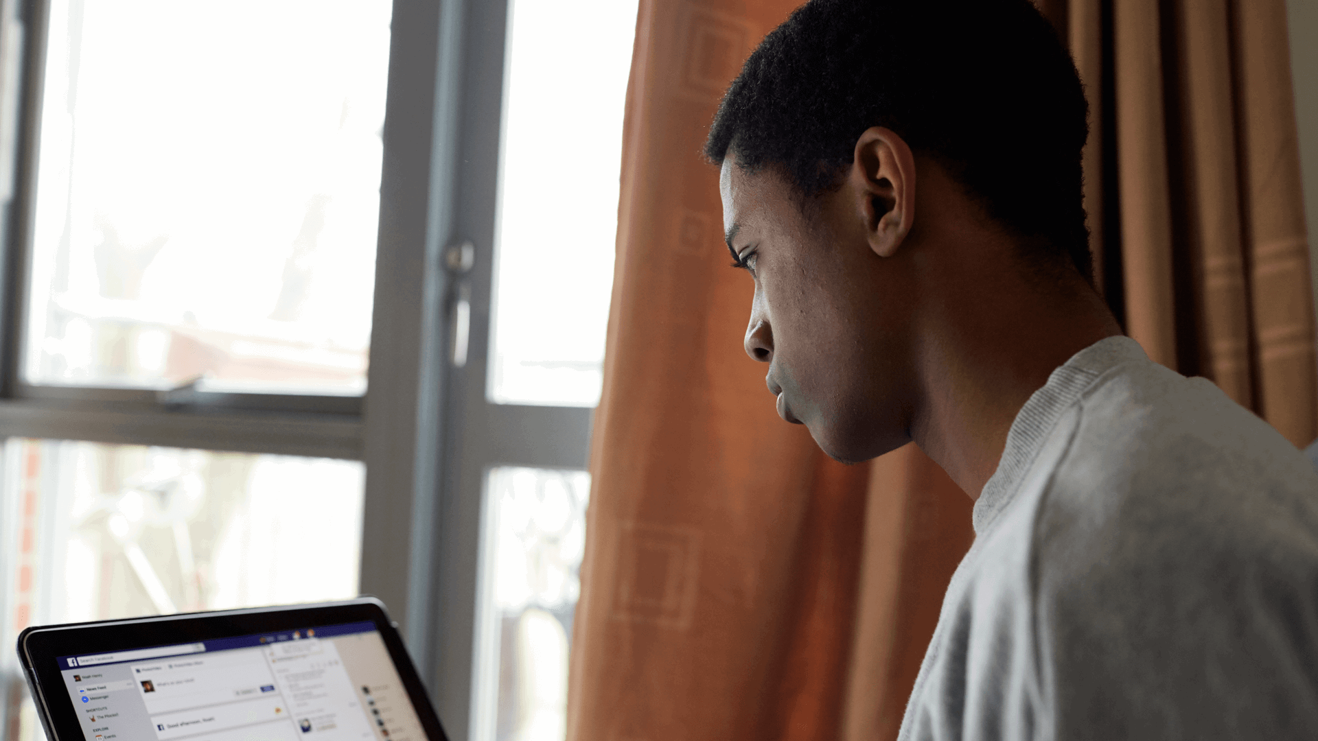 A boy wearing a grey t-shirt sits beside a window while using Facebook on his laptop.