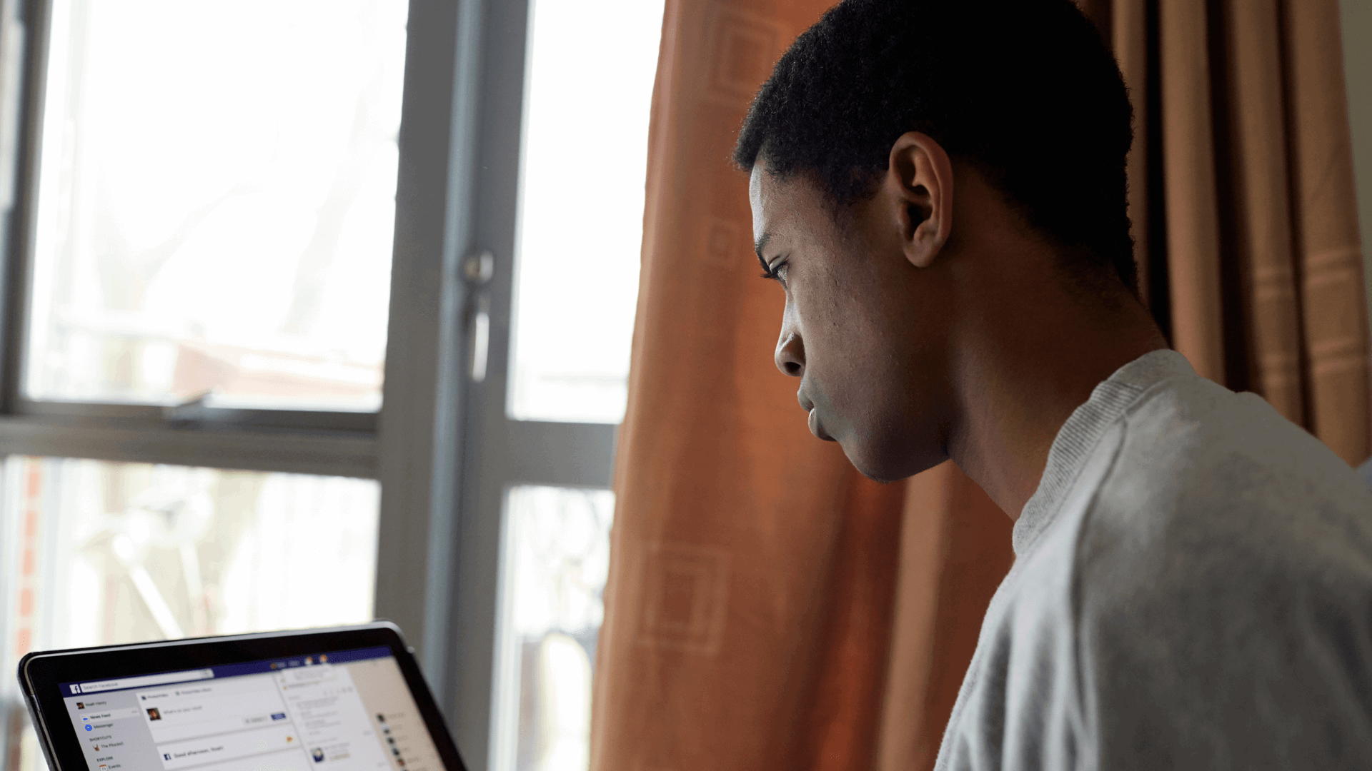 medium shot of a boy wearing white shirt looking a this laptop beside a window