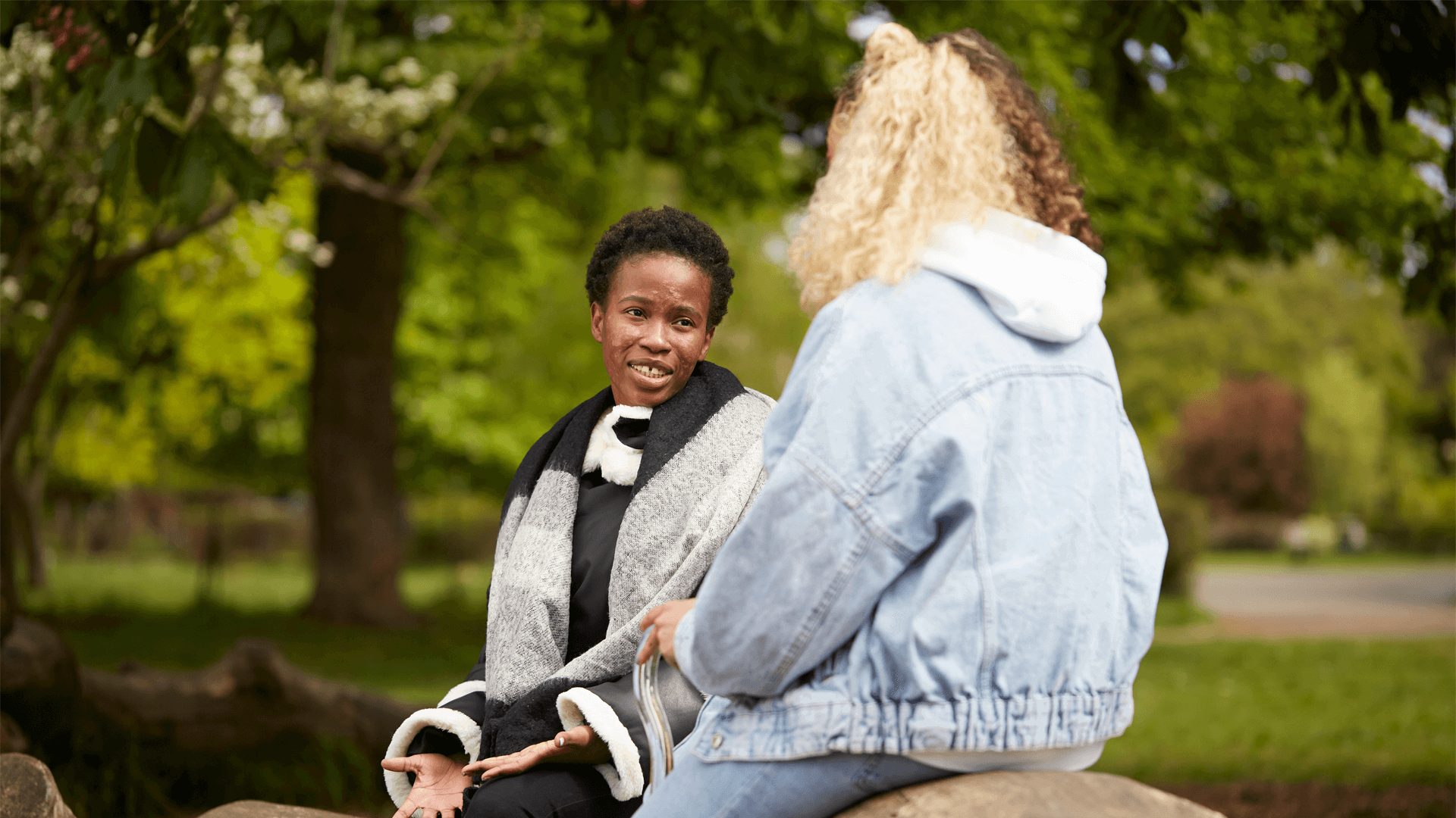 a-girl-with-short-curly-hair-wearing-black-jacket-talking-to-another-young-woman-with-long-curly-hair-wearing-denim-jacket-while-they-are-sitting-on-bench-in-the-park