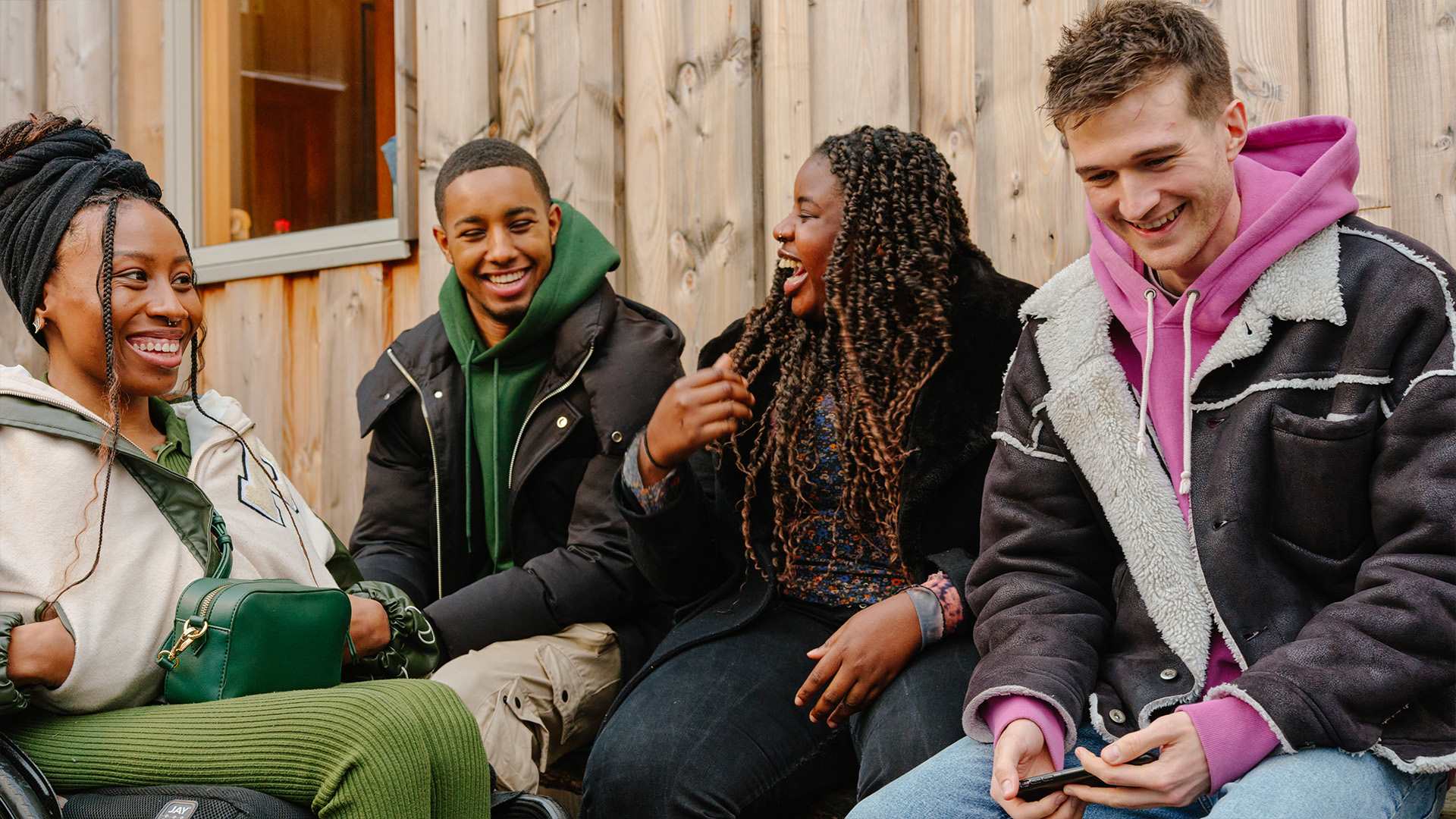 A group of young people laughing together outside on a bench. Group includes two black girls (one in a wheelchair), one black boy, and a white boy.