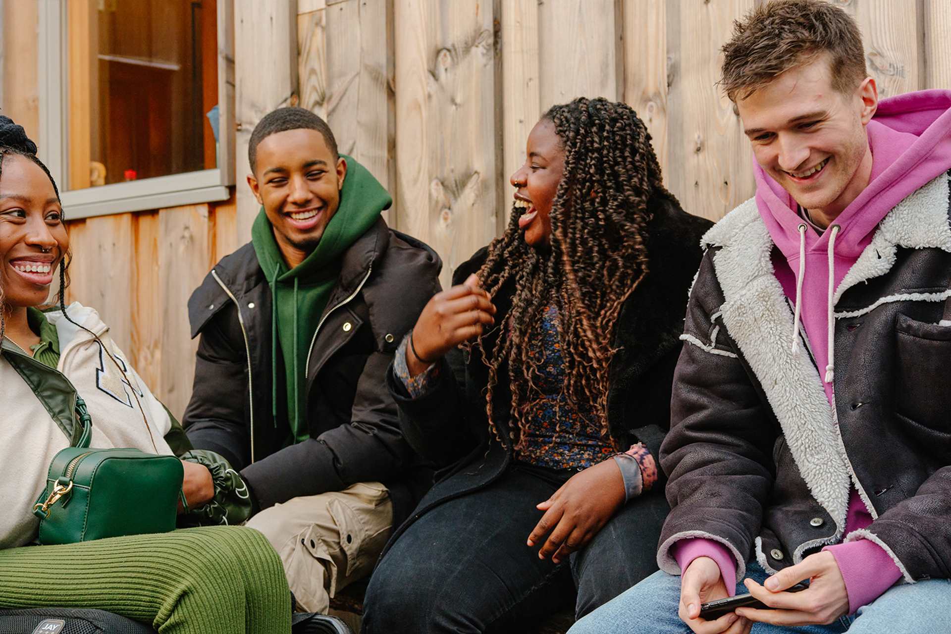 A group of young people laughing together outside on a bench. Group includes two black girls (one in a wheelchair), one black boy, and a white boy.