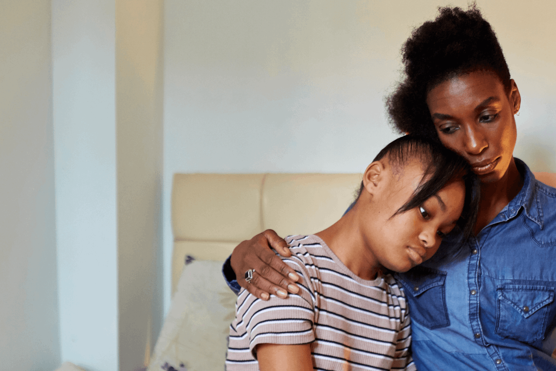 A mother putting her arm around her sad daughter in a bedroom