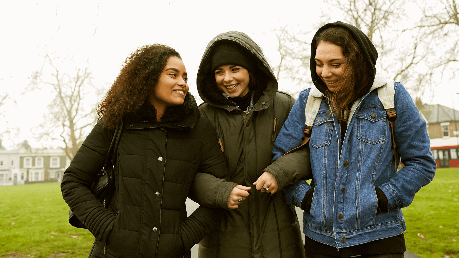 Three young people linking arms and walking through a park together.