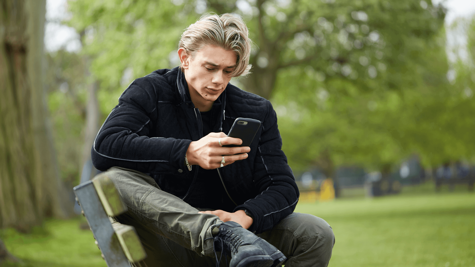 A young man wearing a black jacket sits on a park bench. He is looking at his phone with a worried expression.