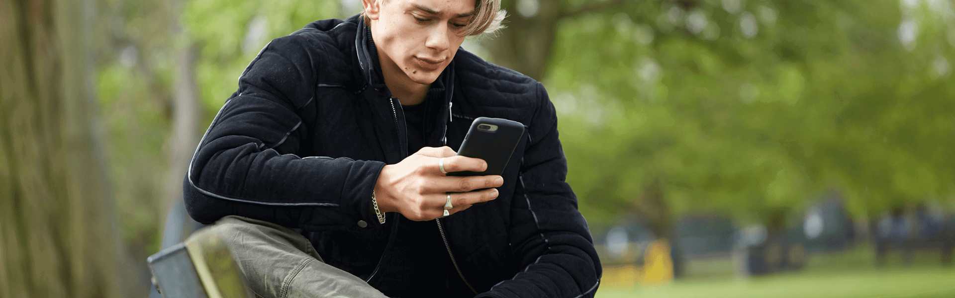 A young man wearing a black jacket sits on a park bench. He is looking at his phone with a worried expression.