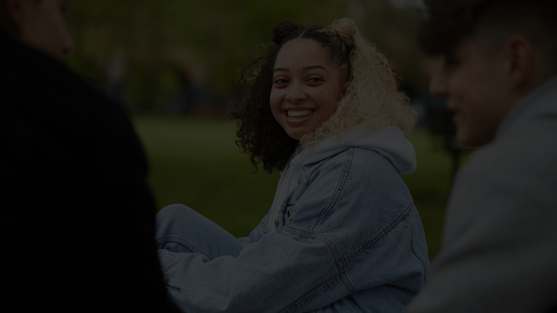 A young woman with curly hair wearing a blue denim jacket is smiling and talking with two young men in the park.