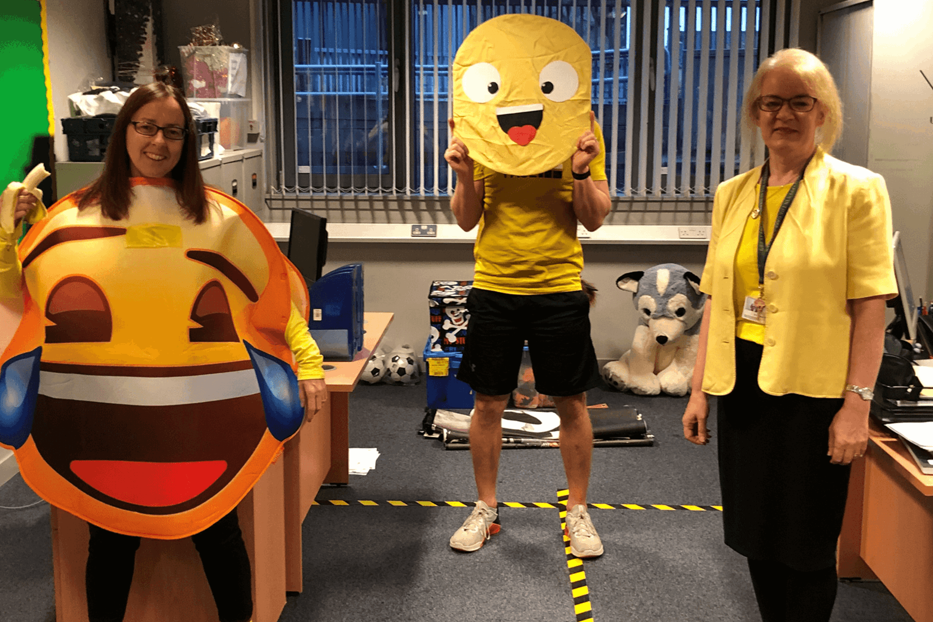 Three people stand in a group wearing yellow, the person on the left is wearing a yellow laughing emoji, the person in the middle holds a yellow smiley face and the person on the right is wearing a yellow top and cardigan