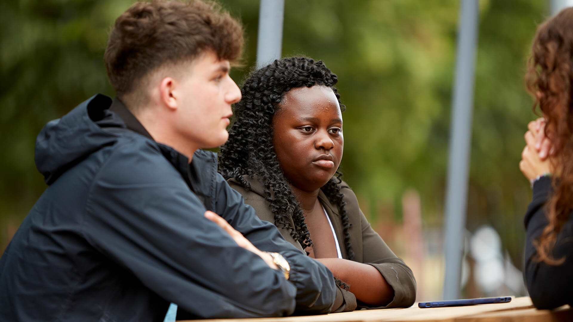 close-up-of-a-young-girl-with-curly-hair-and-a-boy-in-blue-jacket-looking-serious-at-another-girl-cropped-in-the-photo-while-sitting-on-a-bench-with-table-in-the-park