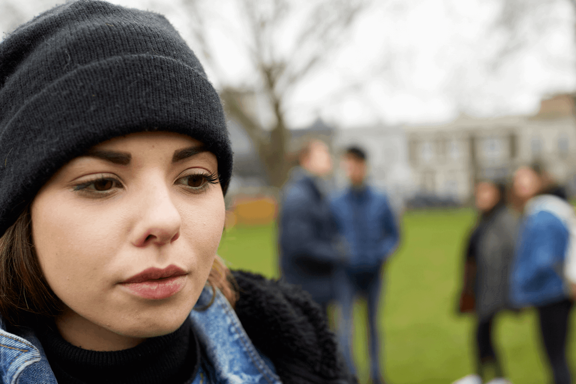 close up of a girl wearing a black beanie feeling left out by friends standing behind her with school and trees on background