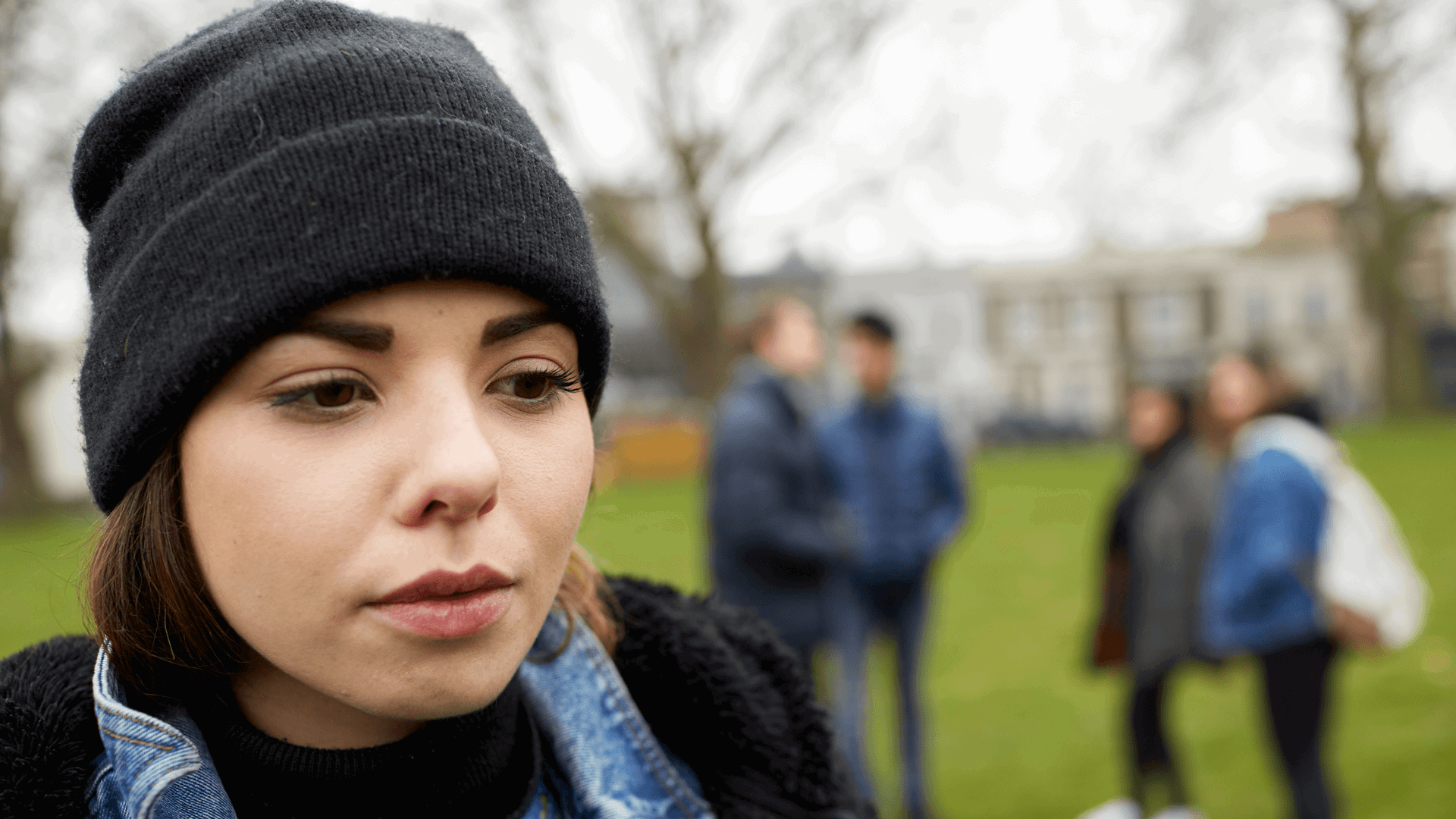 close up of a girl wearing a black beanie feeling left out by friends standing behind her with school and trees on background