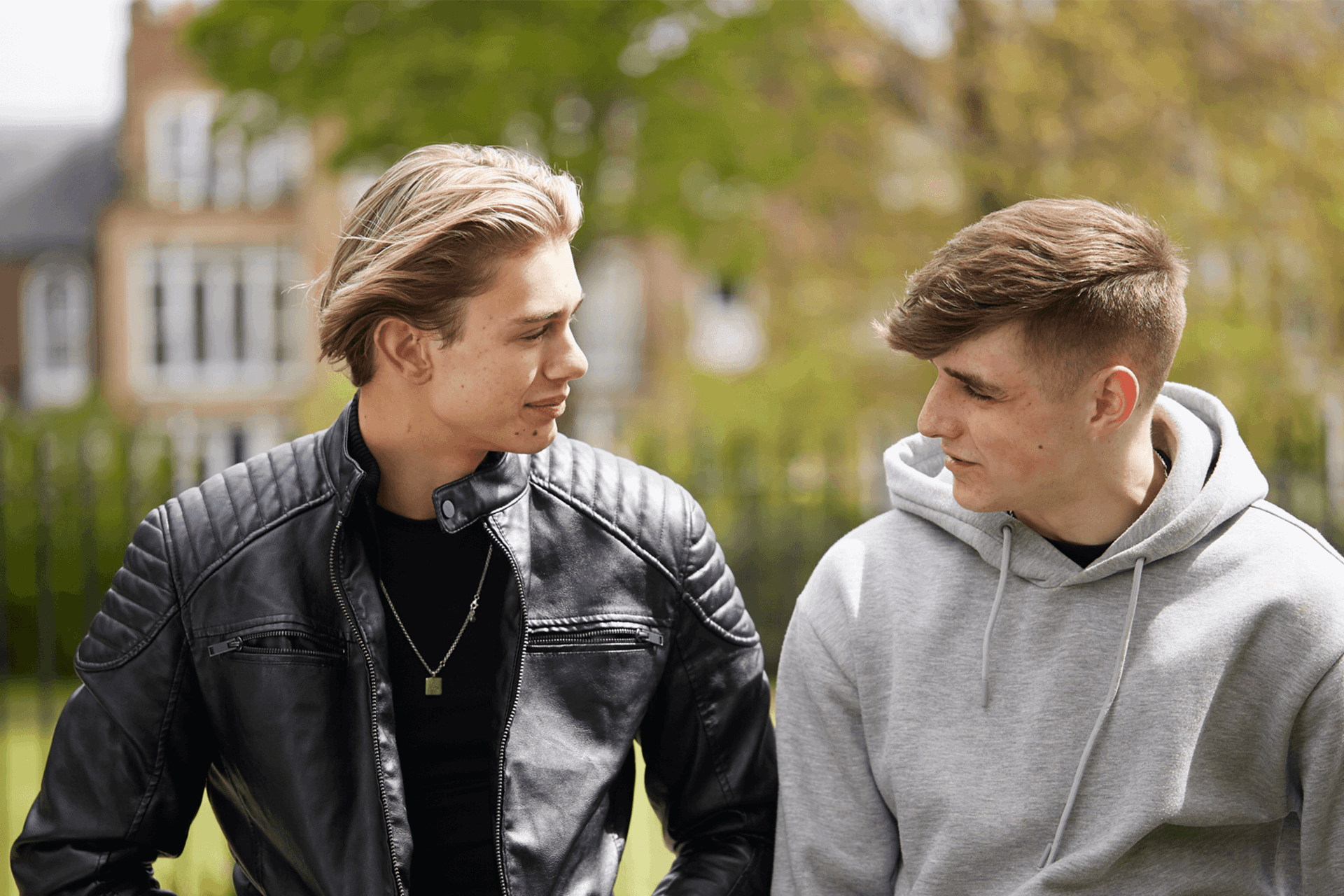 Two boys sitting side-by-side in a park and looking at each other while talking.