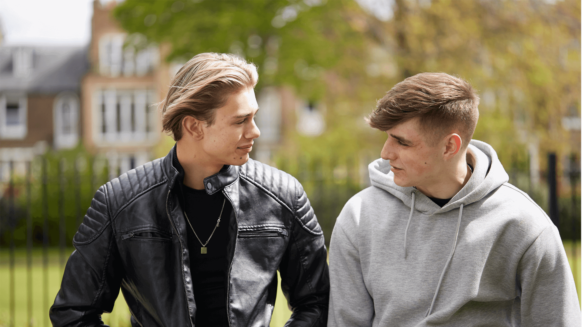 Two boys sitting side-by-side in a park and looking at each other while talking.