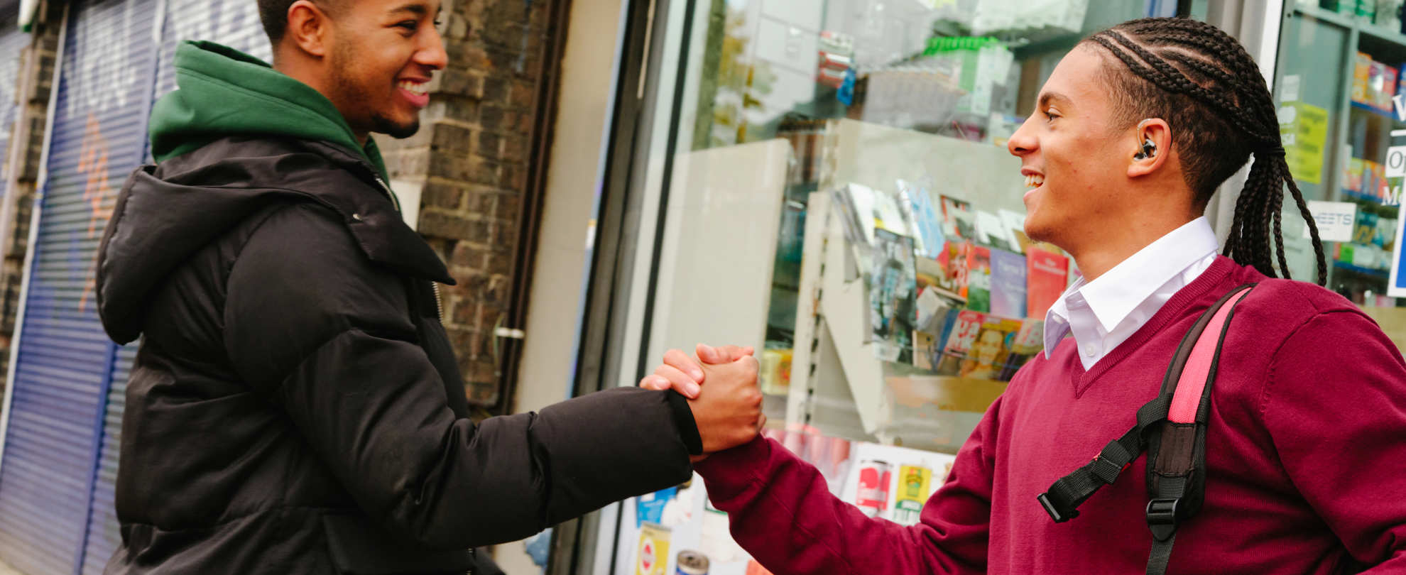 A Black teenage boy with a hearing aid grasping hands with a young Black man. They are standing on the street outside a shop, both smiling.
