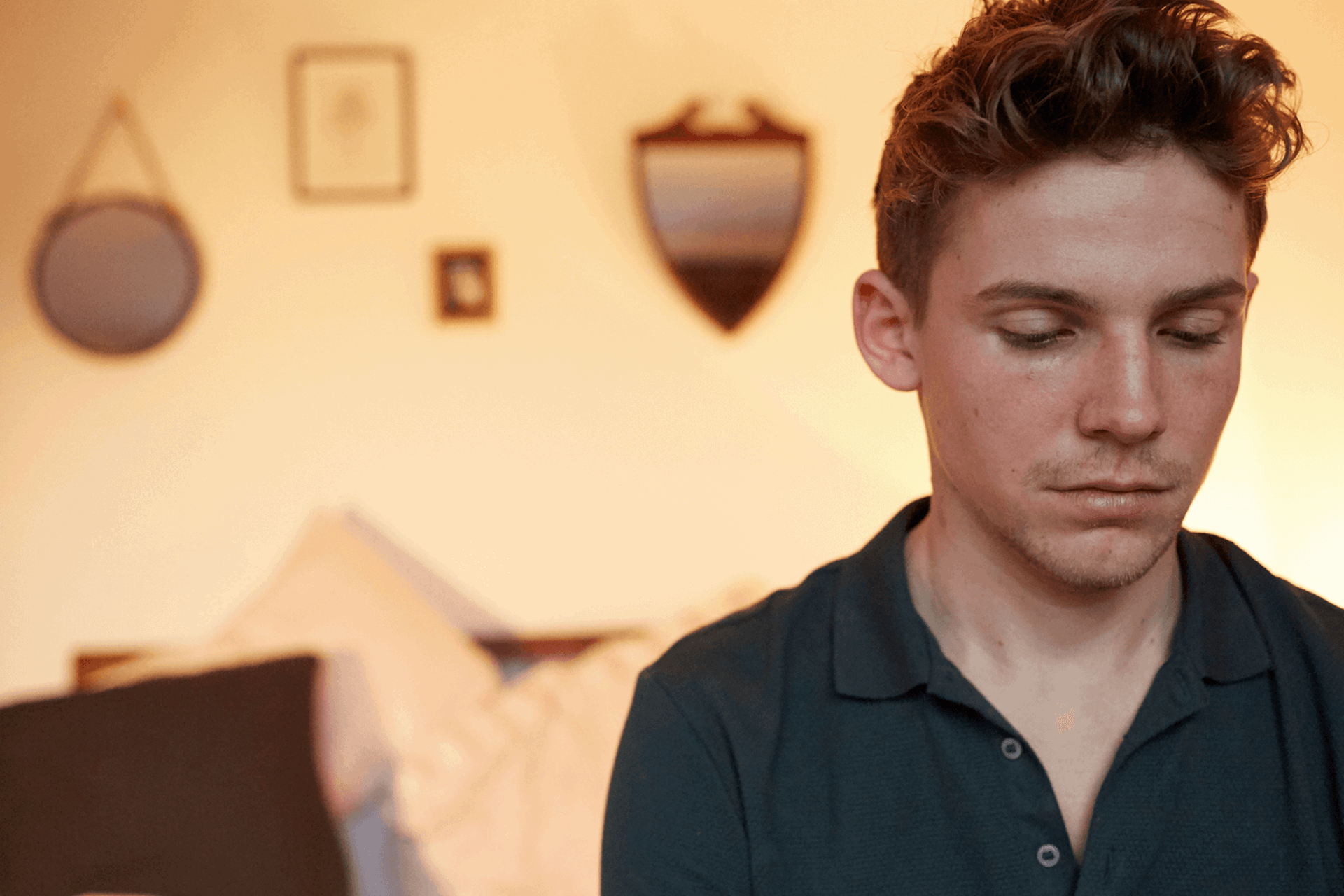 A boy sitting alone in his bedroom. He looks sad with his head and eyes facing down.