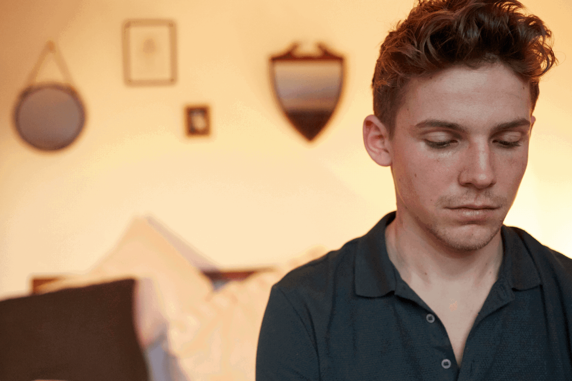 A boy sitting alone in his bedroom. He looks sad with his head and eyes facing down.