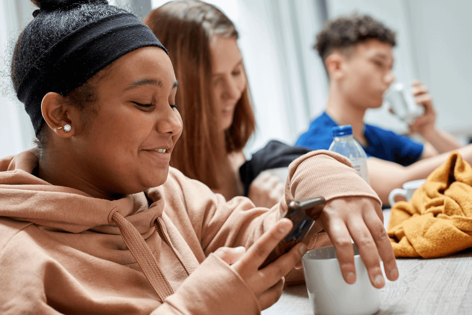 two girls smiling and looking at their phone while sitting on a dining bench having their lunch and a boy drinking water on the background