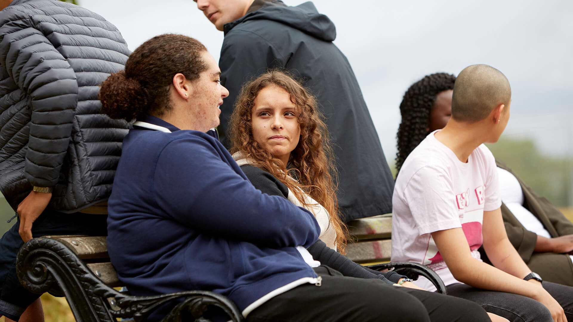 A group of six young people sitting on a bench and talking to each other in a park.