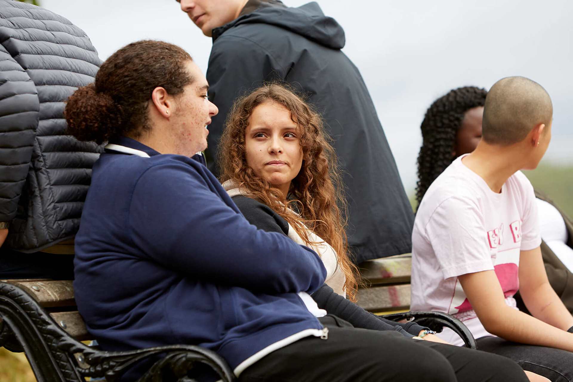 A group of six young people sitting on a bench and talking to each other in a park.