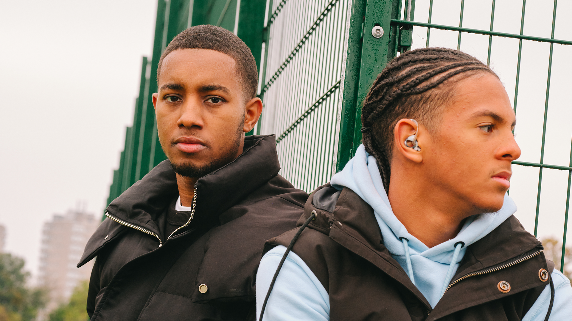 A young Black man sitting in the park with a Black teenage boy wearing a hearing aid. They are both looking very serious.