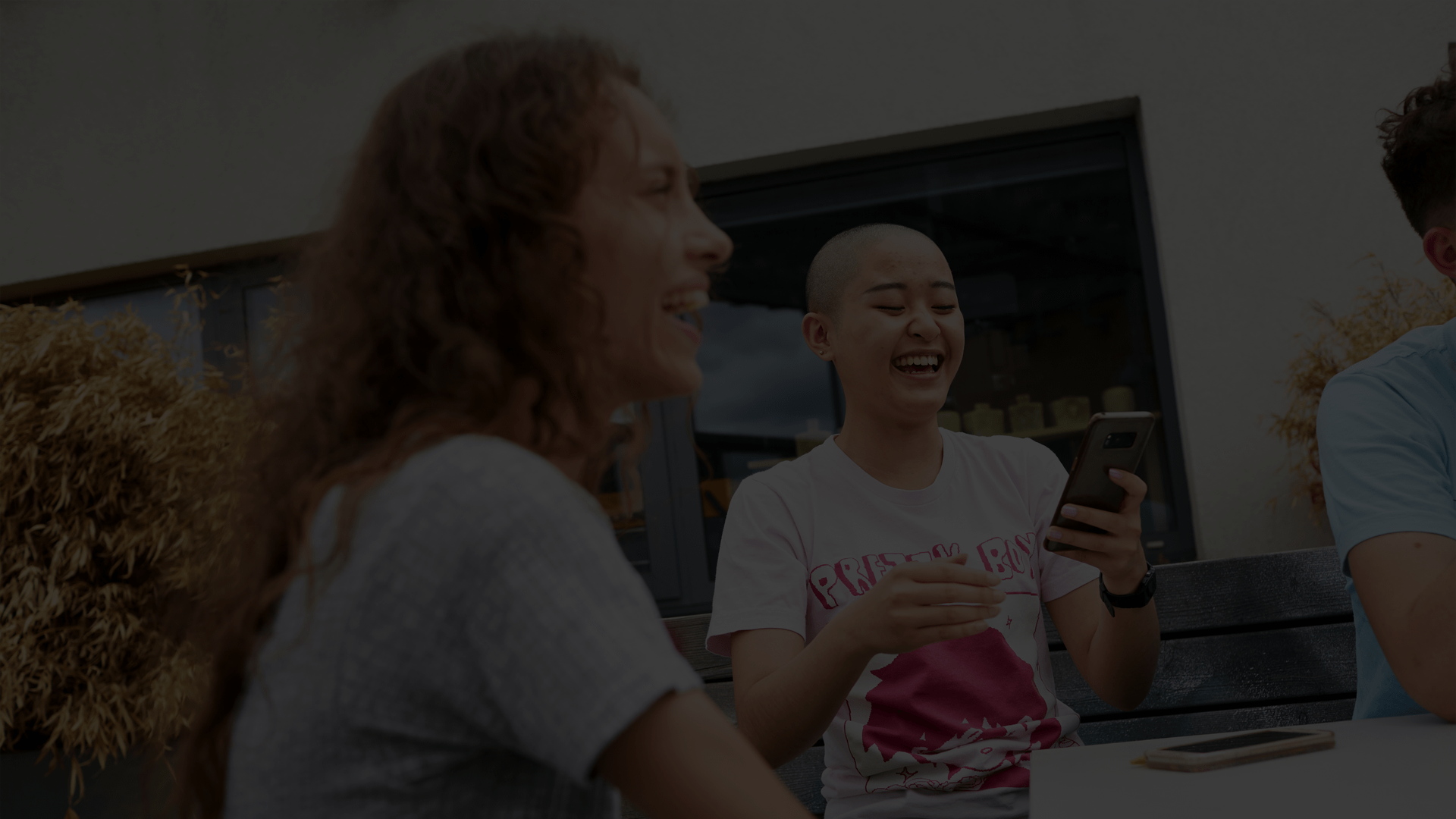 Two girls sitting together at a park bench with their friends. They are both laughing and one is looking at her phone.