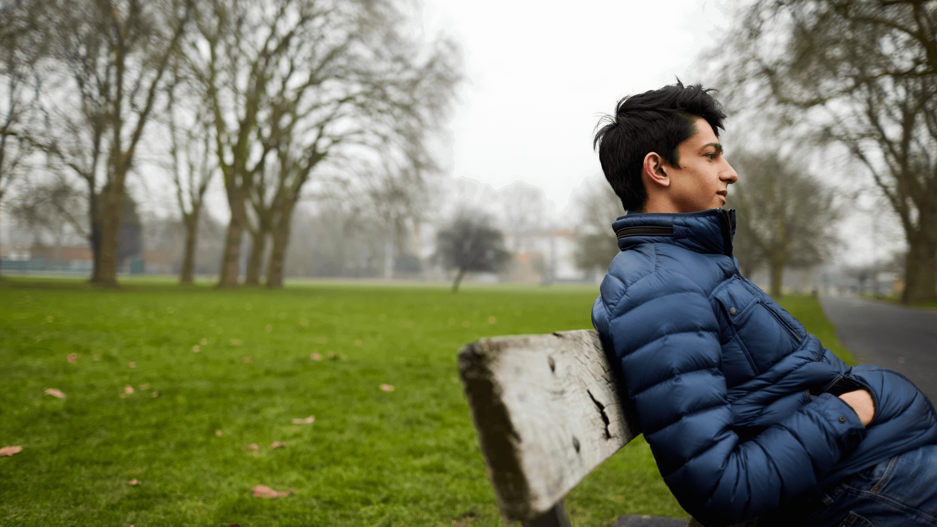 A boy sits on a park bench wearing a blue puffer jacket with his hands in his pockets. He is looking straight ahead as if deep in thought.