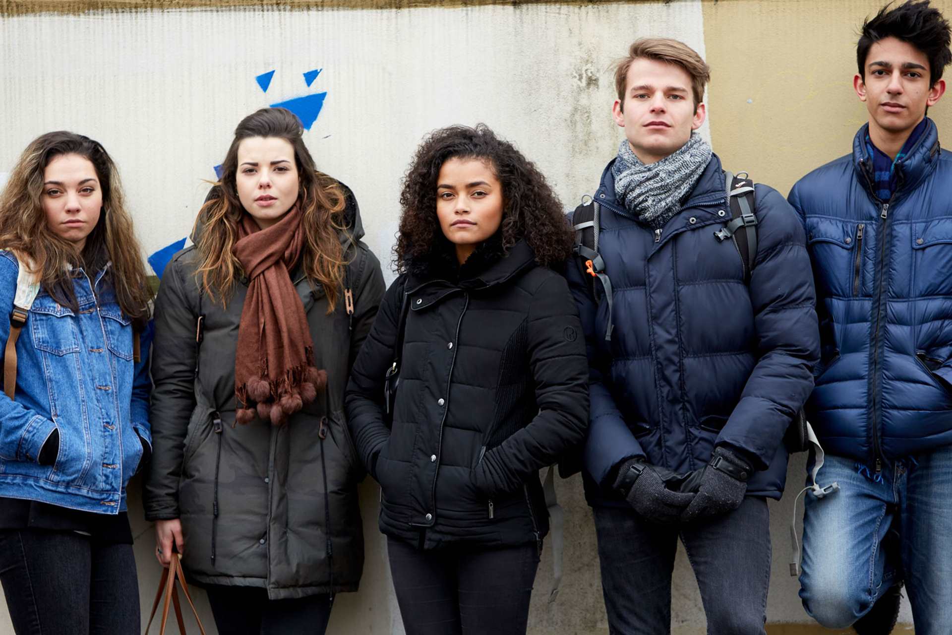 A group of five young people wearing jackets, standing side by side against a wall and looking straight ahead.