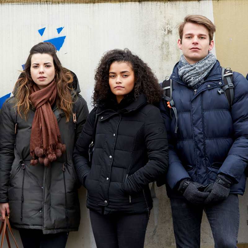 A group of five young people wearing jackets, standing side by side against a wall and looking straight ahead.
