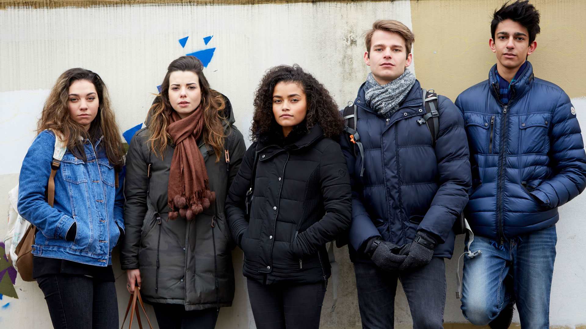 Group of five young people wearing jackets standing against the wall looking in front of the camera.