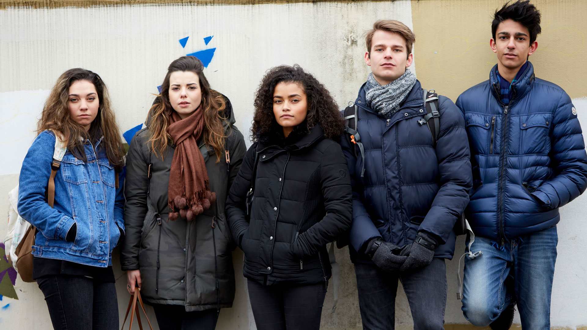 A group of five young people wearing jackets, standing side by side against a wall and looking straight ahead.