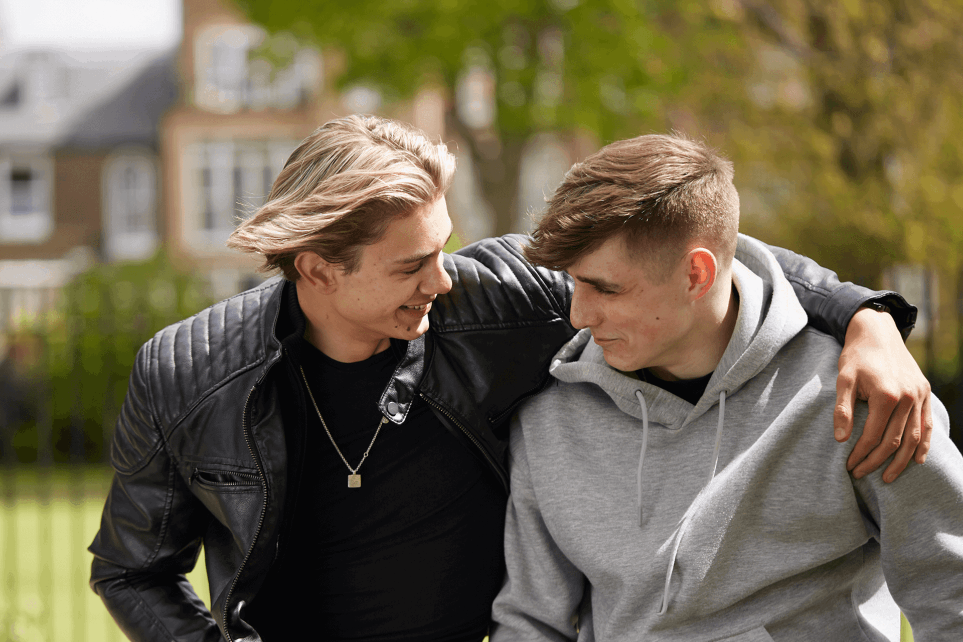 A boy wearing a black jacket is smiling with his arm over his friend's shoulder. They are sitting in a park and smiling at each other.