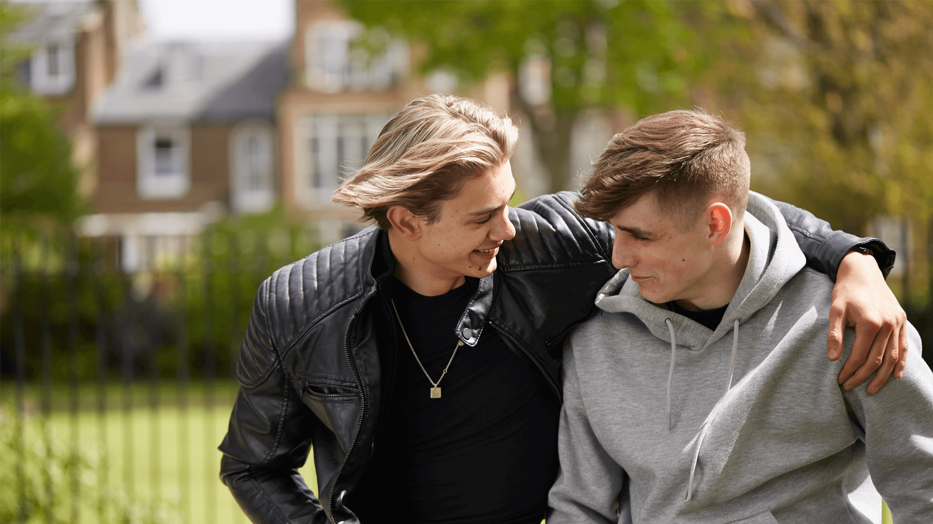 A boy wearing a black jacket is smiling with his arm over his friend's shoulder. They are sitting in a park and smiling at each other.