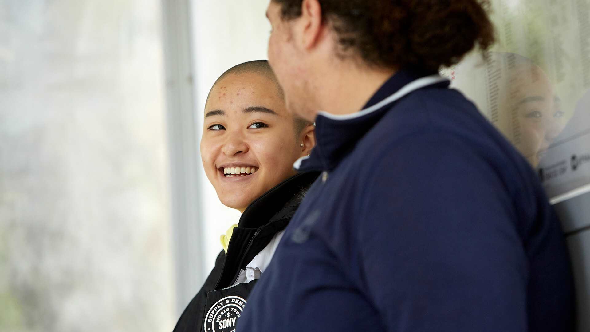 Two young people lean against a bus stop. The person on the left is laughing and wears a black coat while looking at the other young person. The person on the right wears a dark blue jacket and is looking at the person on the left. They're side on to the camera.