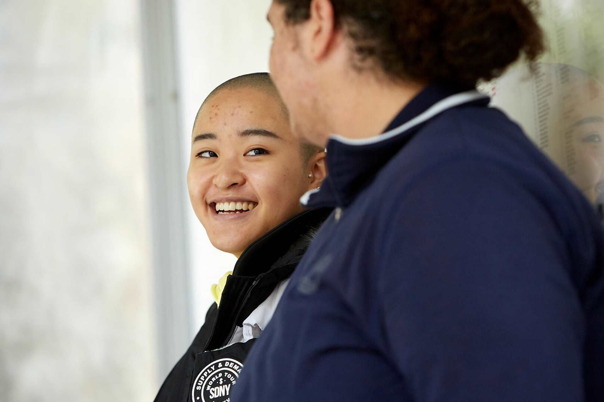 Two young people lean against a bus stop. The person on the left is laughing and wears a black coat while looking at the other young person. The person on the right wears a dark blue jacket and is looking at the person on the left. They're side on to the camera.