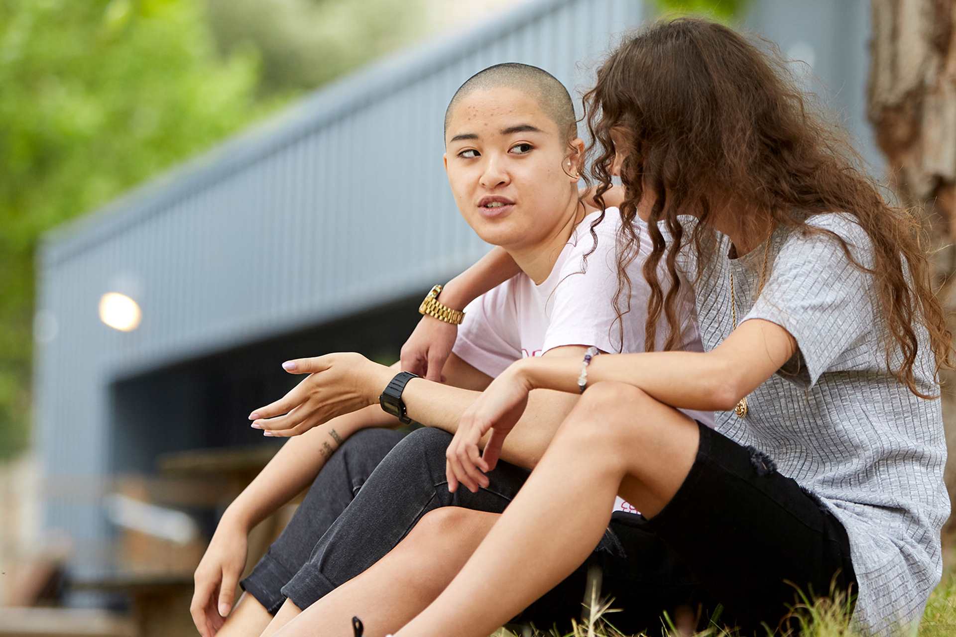 a-young-woman-with-curly-hair-and-in-grey-shirt-and-black-shorts-with-face-unseen-has-her-hands-wrapped-another-young-woman-with-shaved-hair-and-in-white-shirt-and-blakc-trousers-talking-to-her-while-they-sat-on-the-grass-with-a-tree-and-building-on-their-background