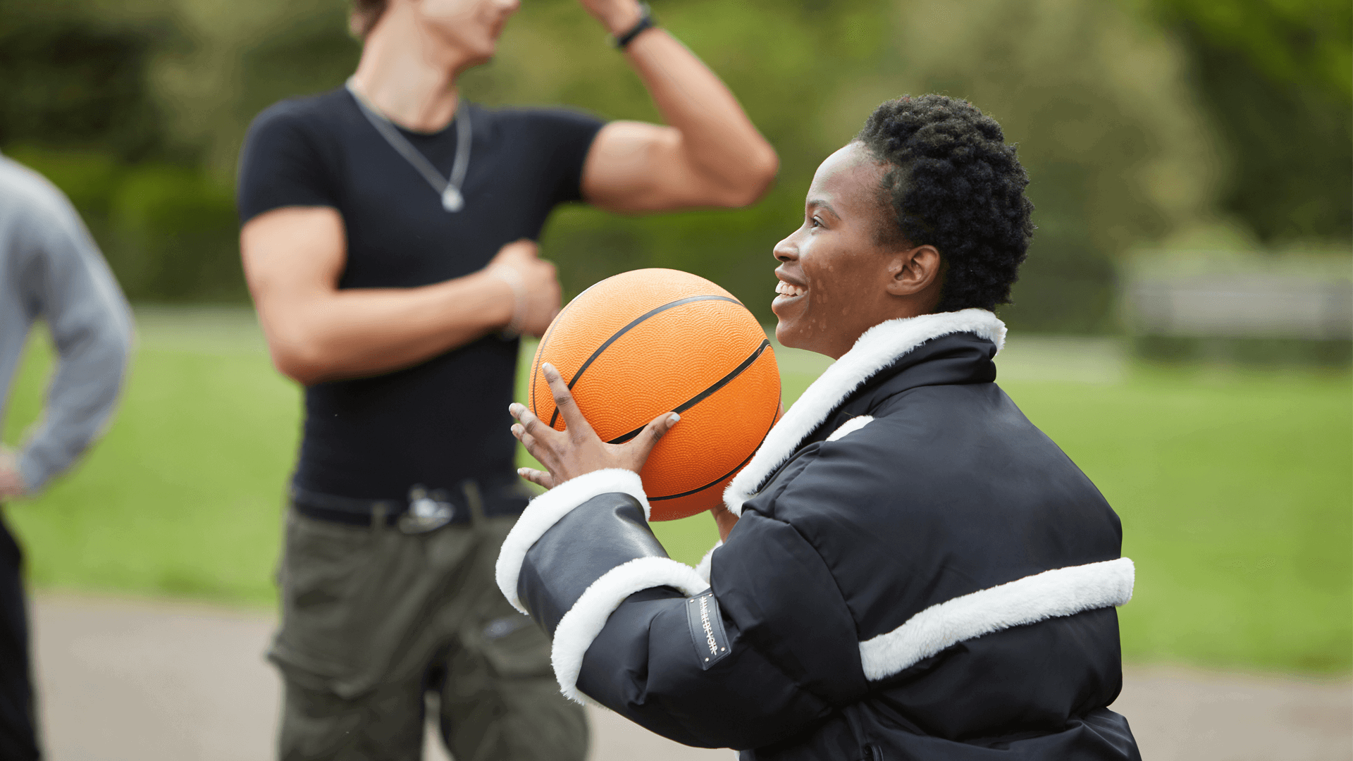 medium shot of a girl with short curly hair wearing black jacket and holding a basketball while smiling and playing with boys