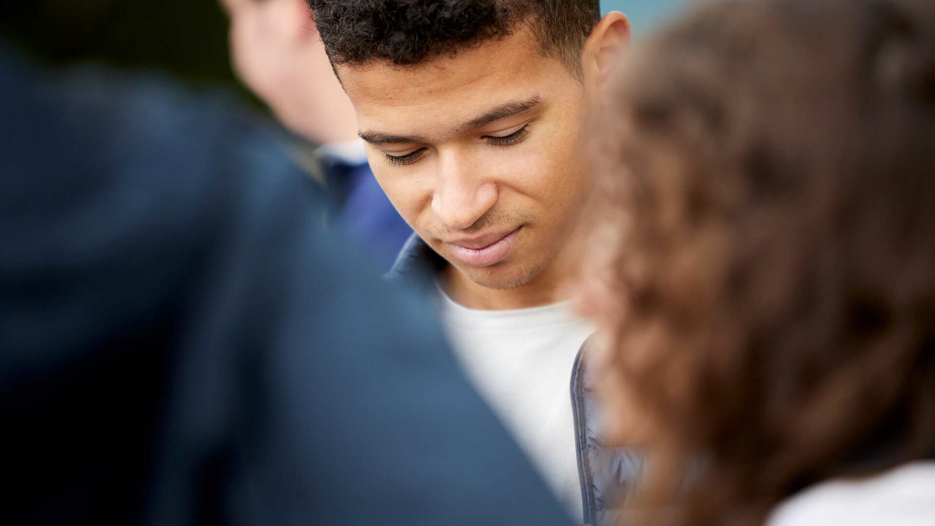 A young man stands in the centre of a group of friends with his eyes and head down.